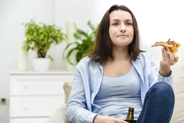 Een jonge vrouw zit op een bank pizza te eten en bier te drinken. Ze steunt haar favoriete team. Lekker-lekker. — Stockfoto