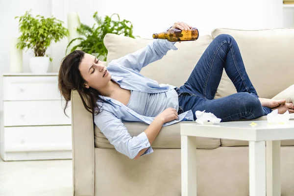 Drunk young woman is lying on the sofa and looking at empty bottle. She wants more.