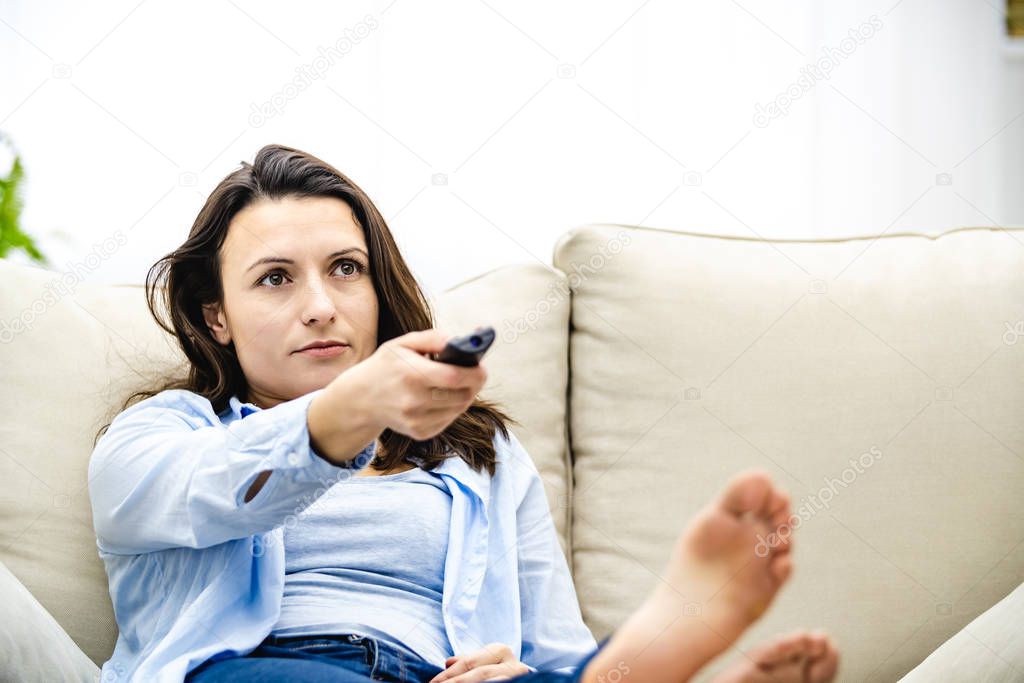 Concentrated young woman is watching tv, laying on a sofa. Copy space.