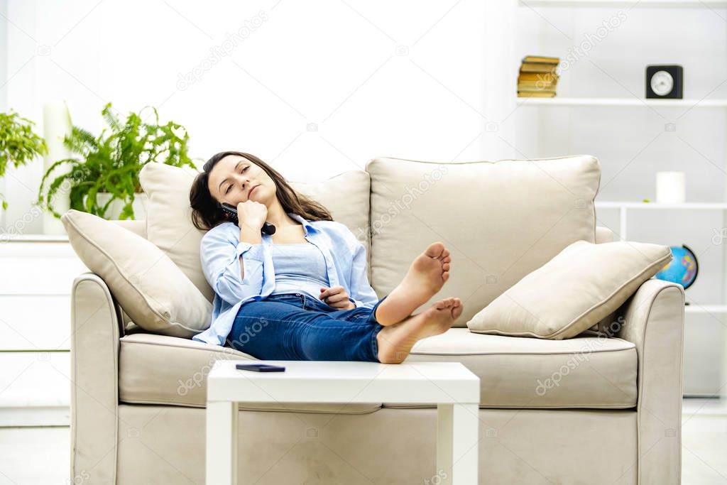 Bored young woman is watching TV, laying on a sofa. Copy space.