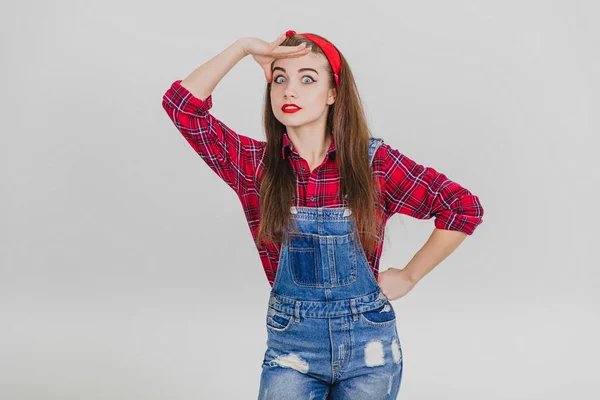 Menina em t-shirt xadrez vermelho e jeans está olhando para a câmera, mantendo a mão perto da testa . — Fotografia de Stock