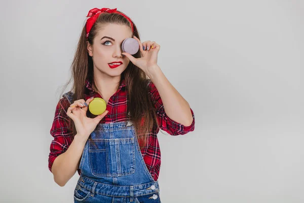 Linda feliz bonito jovem bonita mulher segurando dois macaroons, escondendo seu olho atrás de um azul, torcendo o lábio . — Fotografia de Stock