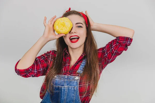Preciosa joven hembra escondiendo su ojo detrás de una gran rosquilla con glaseado amarillo, sonriendo . — Foto de Stock