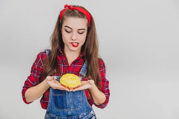 Preciosa joven hembra sosteniendo sabrosa rosquilla grande con glaseado amarillo en sus manos, torciendo su labio tentadoramente . — Foto de Stock