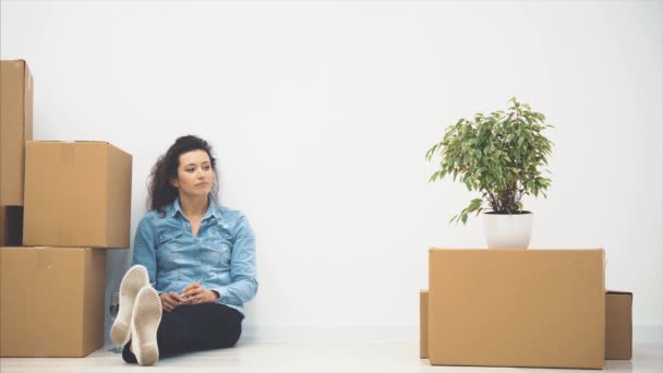 La joven está sentada en el suelo. Hay muchas cajas desnudas y una planta cerca de ella. Copiar espacio. 4K . — Vídeos de Stock