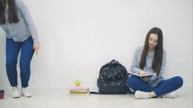 İki sevimli Asyalı kız yerde loto pozisyonunda oturuyor, birbirlerine el sallıyor, parmaklarını uzatıyor, başparmaklarını kaldırıyor, gülümsüyor..