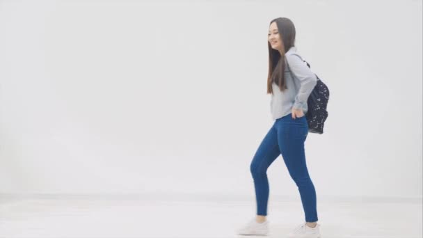 Glad, leende, full höjd asiatisk flicka med långt hår visas på en vit bakgrund med ryggsäck hängande över axeln, händer i bakfickor av hennes jeans, poserar. — Stockvideo