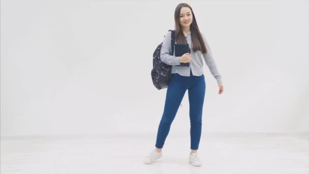 Una colegiala asiática que aparece sobre un fondo blanco. Con una mochila. Mantiene el libro y sonríe . — Vídeos de Stock