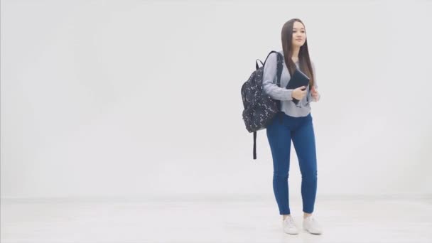 Uma estudante asiática aparecendo em um fundo branco com uma mochila, abrindo livro preto, olhando para ele, fechando, posando . — Vídeo de Stock