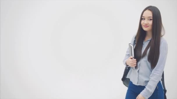 Egy ázsiai iskoláslány jelenik meg fehér háttérben, hátizsákkal a vállán, könyvet tart a mellkasa mellett, pózol.. — Stock videók
