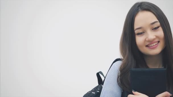 An asian schoolgirl appearing on a white background with a backpack slung over her shoulder, holding book near chest, posing and smiling. — Stock Video