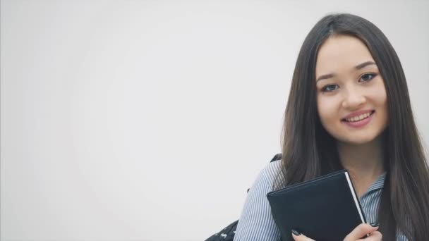 Sırtında sırt çantasıyla beyaz bir arka planda beliren Asyalı bir kız çocuğu, kitabı göğsüne yakın tutuyor, poz veriyor ve gülümsüyor.. — Stok video