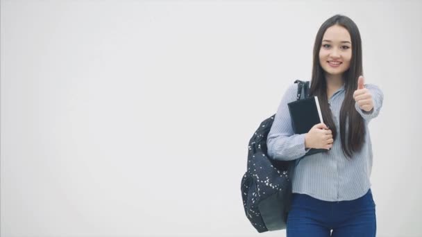 Une écolière asiatique debout avec un sac à dos pendu sur son épaule, tenant un livre près de la poitrine, donnant pouces vers le haut . — Video