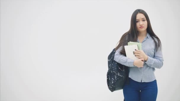 En asiatisk skolflicka visas på en vit bakgrund med en ryggsäck hängde över axeln, hålla en bunt böcker nära bröstet, suckar, ser trött. — Stockvideo
