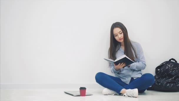 Jovem menina asiática envolvida no processo de leitura de livro e olhando ao redor enquanto sentado no chão em lotos posição . — Vídeo de Stock