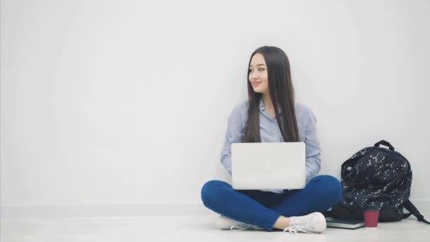 Kedves fiatal ázsiai hölgy ül a padlón lótusz pozícióban, laptoppal, oldalra néz, felemeli a hüvelykujját, mosolyog. — Stock videók