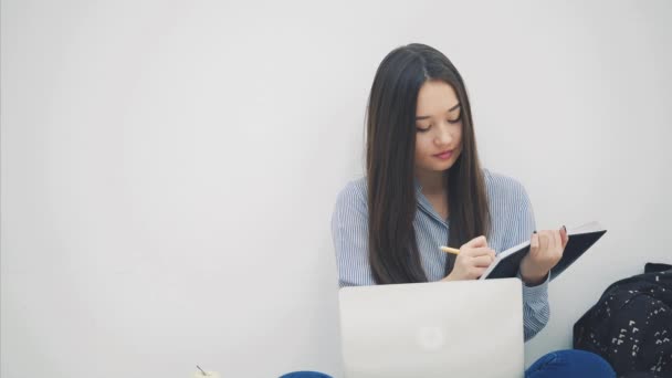 Asiática dama sentada en el suelo en posición lotos, con portátil, copiando algo en su cuaderno . — Vídeos de Stock