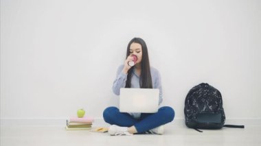 Gülümseyen Asyalı kız öğrenci loto pozisyonunda oturuyor, dizüstü bilgisayar kullanıyor, kahve içiyor..
