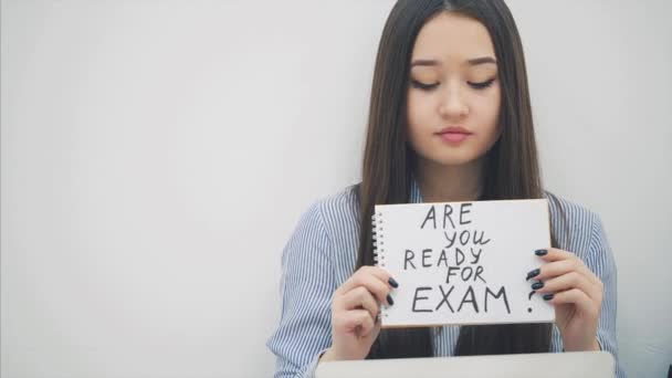 Schöne junge asiatische Dame sitzt, mit Laptop, hebt ein Blatt Papier mit "Sind Sie bereit für die Prüfung" darauf geschrieben, seufzt schwer. Fröhlicher Gesichtsausdruck. — Stockvideo