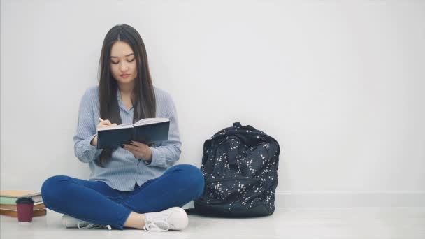 Inspired young asian girl sitting on the floor, finished writing, took out phone and headphones out of her backpack, and is going to listen to music. — ストック動画