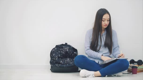 İlham verici Asyalı genç bir kız yerde oturuyor, yazmayı bitiriyor, telefon ve kulaklık alıyor ve müzik dinliyor, dans ediyor.. — Stok video