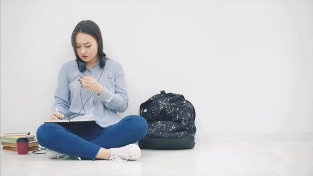 Gadis Asia muda duduk di lantai, bosan menulis, menguap. Jadi, dia mengambil telepon dan headphone dan mendengarkan musik, menari . — Stok Video