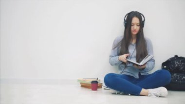 Komik genç Asyalı kız yerde kulaklıkla oturmuş, müzik dinliyor, yazmayı bırakıyor ve selfie çekiyor, poz veriyor, barış işareti gösteriyor..