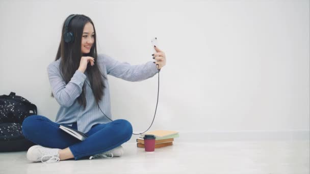 Funny young asian girl sitting on the floor in headphones, listening to the music, takes selfie, posing, waving hello, then messaging with someone. — Stock Video
