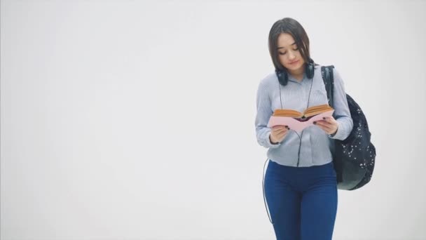 En asiatisk skolflicka med en ryggsäck framträder på en vit bakgrund, läser en bok, suckar, ser olycklig. — Stockvideo