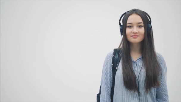 戴着耳机的年轻漂亮的亚洲女孩披着背包挂在肩上，对着相机飞吻了一下，微笑着. — 图库视频影像