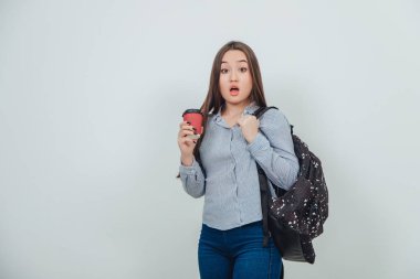 Sırt çantasıyla elinde bir fincan kahveyle kameraya bakıp hayretler içinde bakan genç ve güzel bir kız öğrenci..