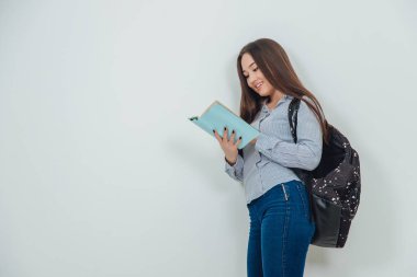 Elinde mavi kitapla dikilen sevimli Asyalı genç bir kız okuma sürecinde yer alıyor. Sırt çantası omzuna asılmış..