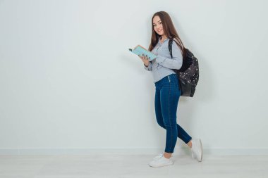 Asyalı tatlı bir kız ayakta, interdting kitabı okuyor. Sırt çantası omzuna asılmış..