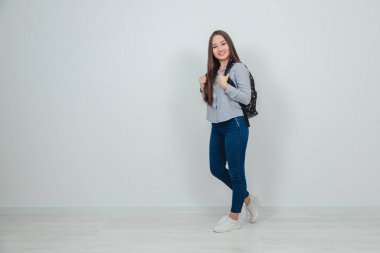 Güzel gülümseyen Asyalı genç liseli kız kot pantolon ve gömlek giyiyor, okul sırt çantasıyla omuzlarının arkasında. İlham verici görünüş.