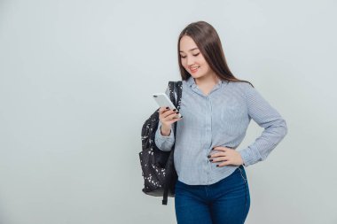 Genç Asyalı öğrenci ayakta duruyor, telefona bakıyor, el akimbo, sırt çantası omzunda asılı..