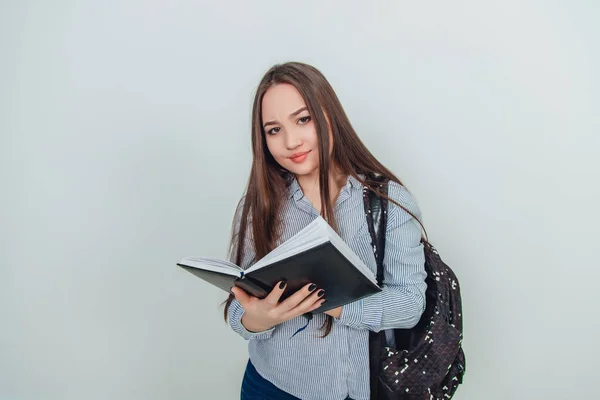 Cute młoda Azjatka dziewczyna stoi, trzymając czarny notatnik otwarty, patrząc ciekawie w aparacie. Plecak zawieszony na jej ramieniu. — Zdjęcie stockowe