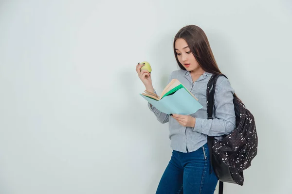 Roztomilá mladá Asiatka stojí, čte zajímavou knihu, drží žluté jablko. Batoh přehozený přes rameno. — Stock fotografie