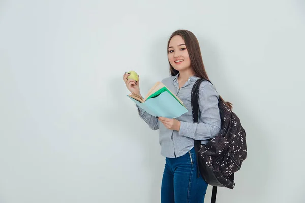 Roztomilá mladá Asiatka stojí, drží zajímavou knihu a žluté jablko, dívá se do kamery, usmívá se. Batoh přehozený přes rameno. — Stock fotografie