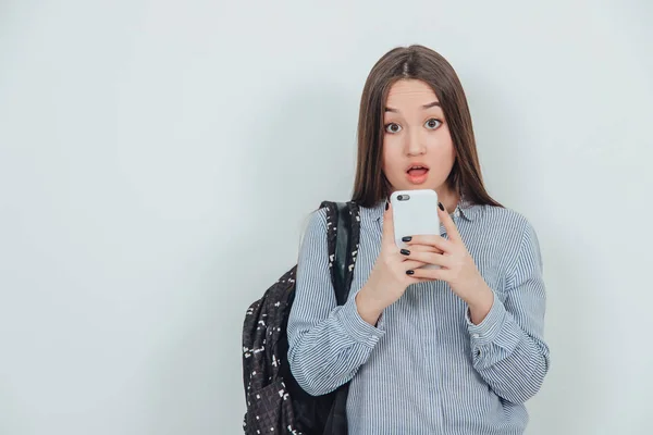 Aranyos ázsiai diáklány, kezében egy telefonnal, mintha fotózna, pózolna. Ruksack rávetette magát a vállára. Kifejező arc. — Stock Fotó