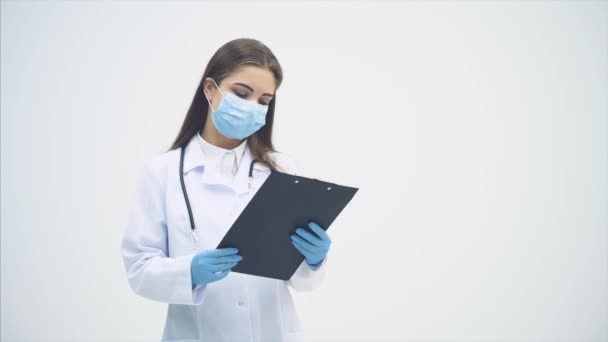 Jovem estagiária em pé de uniforme médico e máscara, segurando prancheta, verificando dados, sacudindo a cabeça, virando páginas . — Vídeo de Stock