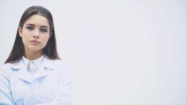 Jovem estagiária em pé de uniforme médico, olhando seriamente para a câmera, posando, com as mãos cruzadas . — Vídeo de Stock