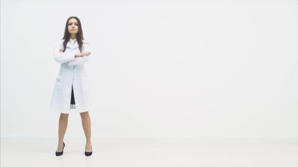 Полноразмерная молодая женщина-врач, стоящая в медицинской форме, глядя на камеру с концентрированным выражением лица, позируя, сложив руки . — стоковое видео