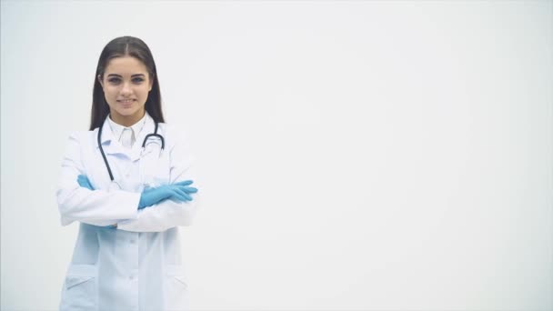 Jovem médica aparecendo no fundo branco, olhando para a câmera, sorrindo, posando, com as mãos dobradas . — Vídeo de Stock