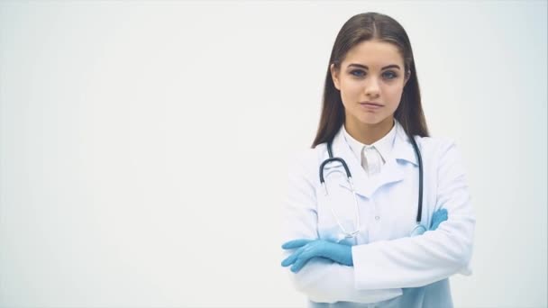 Jovem médica aparecendo no fundo branco, olhando para a câmera, sorrindo, posando, com as mãos dobradas . — Vídeo de Stock