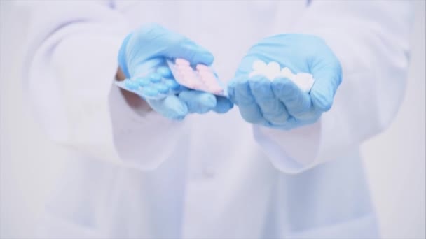 Muchas pequeñas pastillas blancas y dos blísteres de píldoras azules y rosas en las manos de los médicos vestidos con los guantes médicos desechables . — Vídeos de Stock