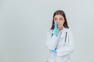 Genç bir kadın stajyer ayakta, tıp üniforması giyiyor, kameraya bakıyor, sanki birinden saklanıyormuş gibi fısıldıyor..