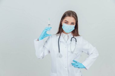 Tıbbi maskeli ciddi bir kadın doktor elinde şırınga, elinde akimbo.