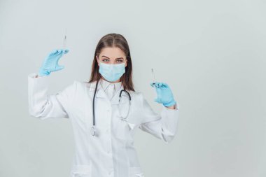 Kendine güvenen, ciddi, tıbbi maskeli bir kadın doktor iki şırınga kaldırıyor..