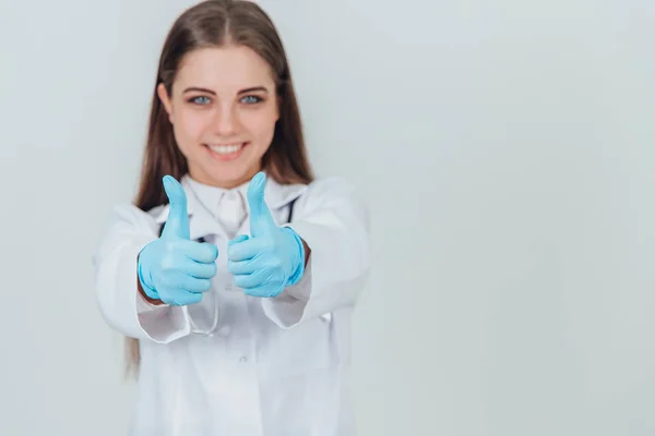 Inspirado jovem praticante com grandes olhos azuis, de pé, vestindo uniforme médico, estetoscópio sobre o pescoço, sorrindo para a câmera, dando polegares para cima . — Fotografia de Stock