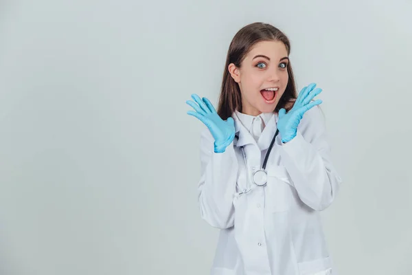 Funny female intern standing in medical uniform, raising her hands, looking at the camera with surprise and satisfaction on her face, mouth and eyes widely opened. — ストック写真